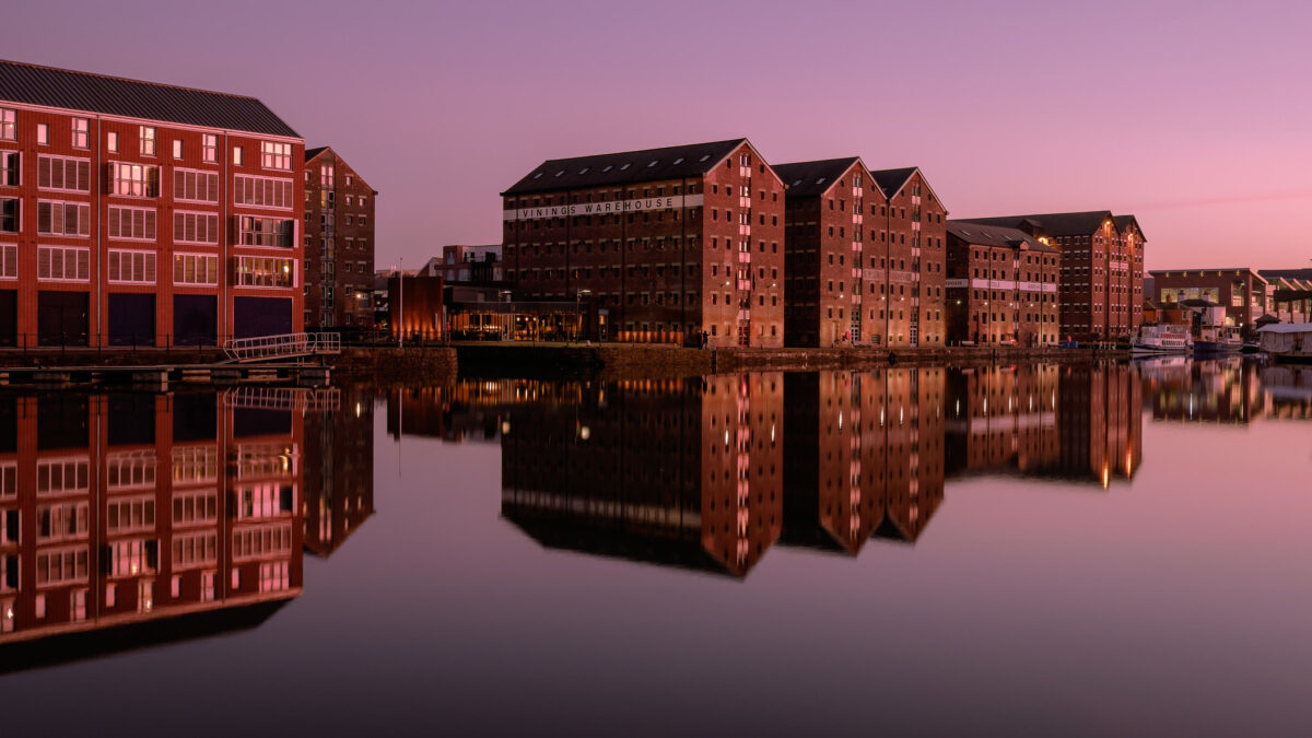 Purple skies over Gloucester docks reflecting off calm water in the foreground with warehouses in the background